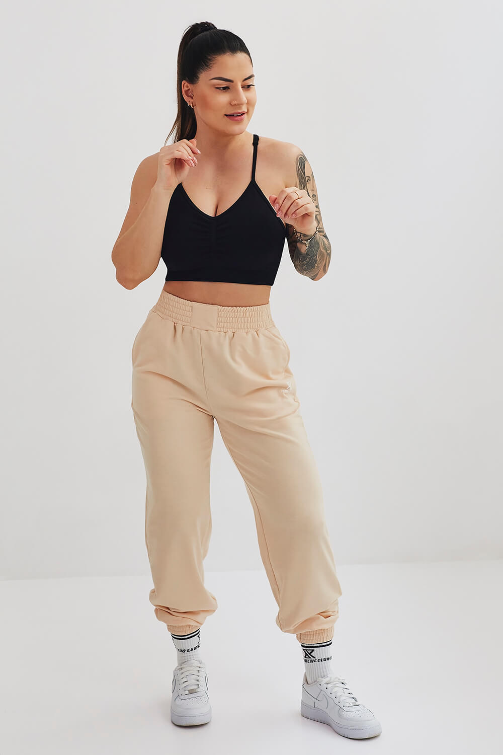 Femme en brassière noire et pantalon de jogging beige, posant en mouvement dans un studio de photographie