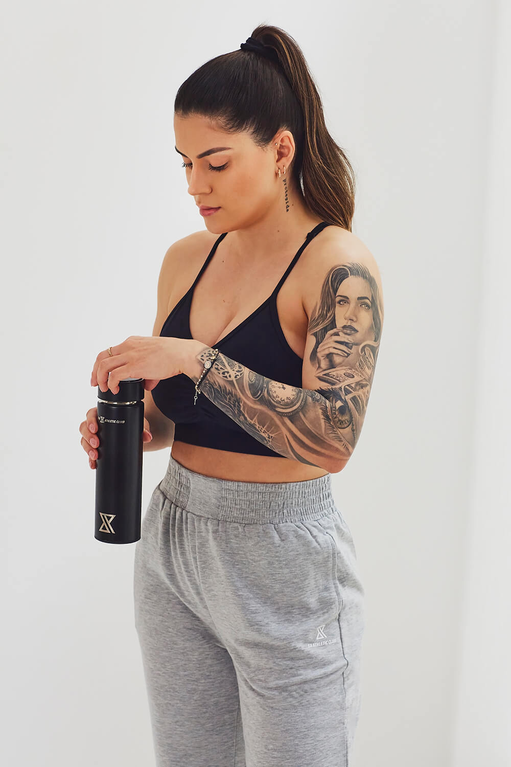 Femme en brassière noire et jogging gris, tenant une gourde de sport noire