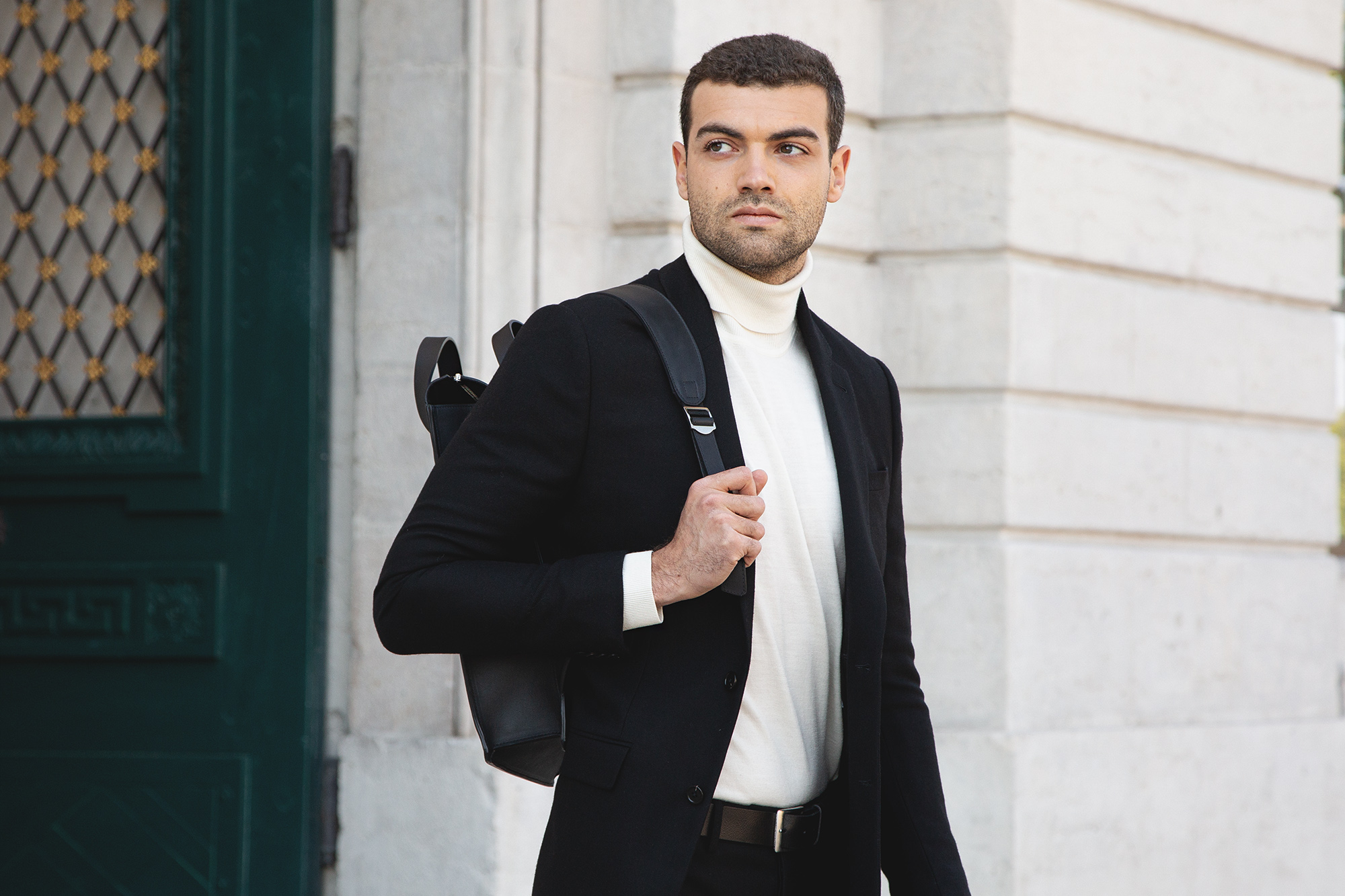 Homme en costume noir avec col roulé blanc et sac à dos, marchant en extérieur
